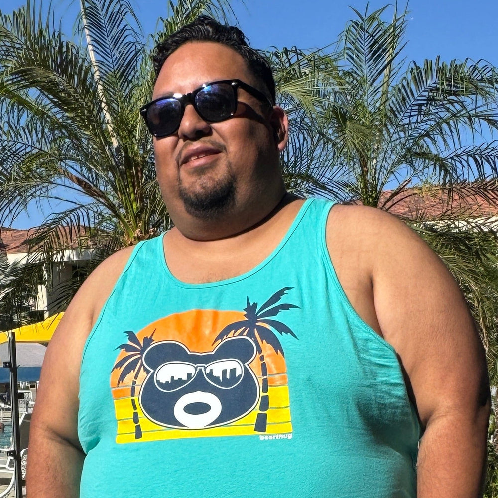 Man wearing a turquoise tank top with a graphic design, sunglasses, and shorts standing on a sidewalk with palm trees and buildings in the background.
